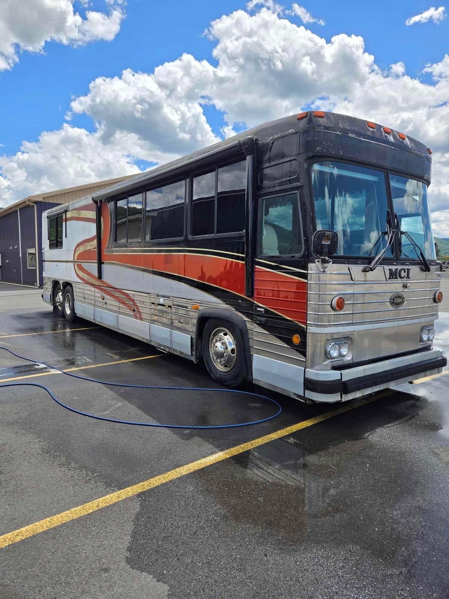RV bus, silver, orange, black paint job, parked on wet asphalt, under cloudy sky.