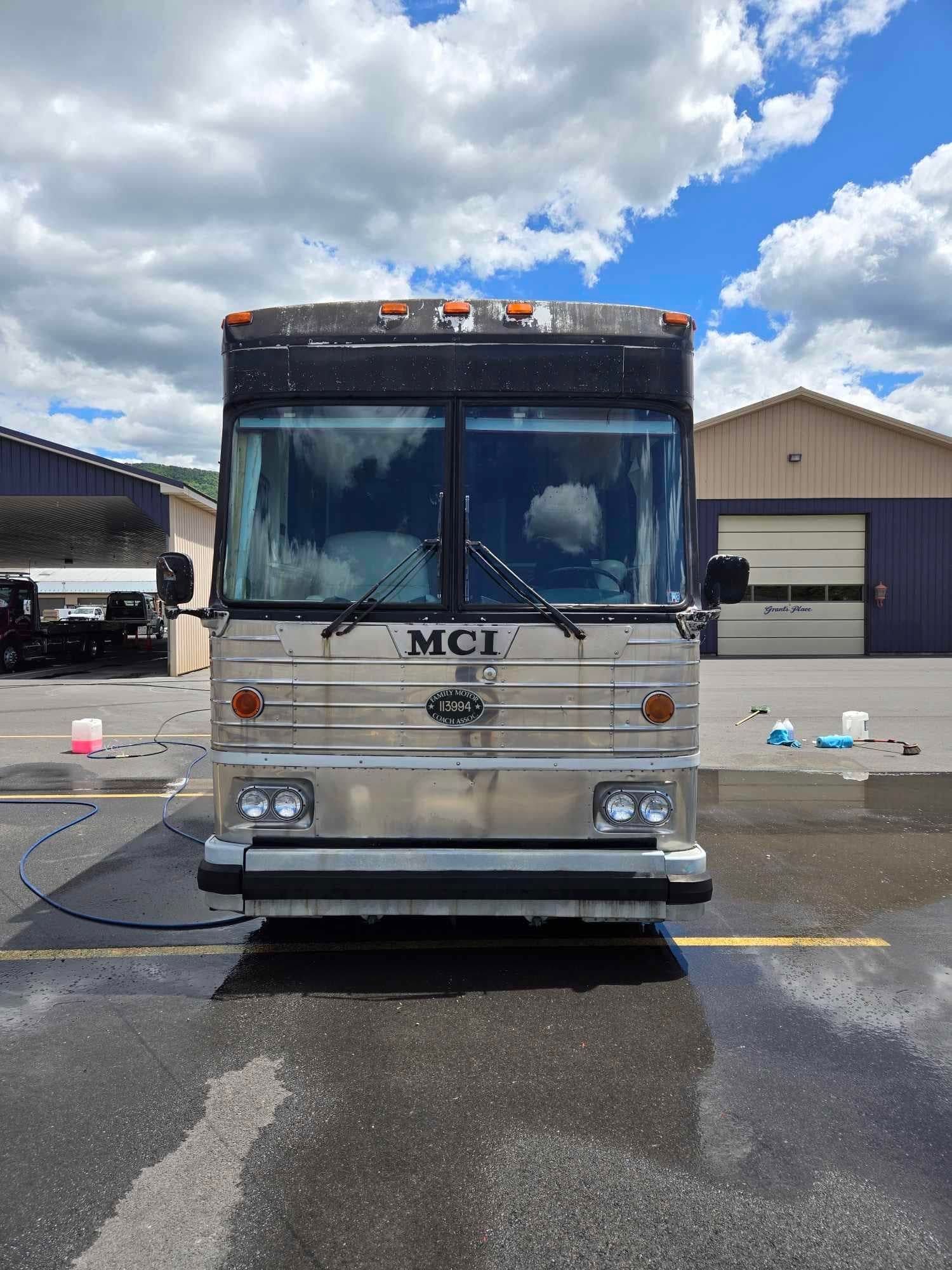 Silver MCI bus being washed outdoors on a bright, partly cloudy day.