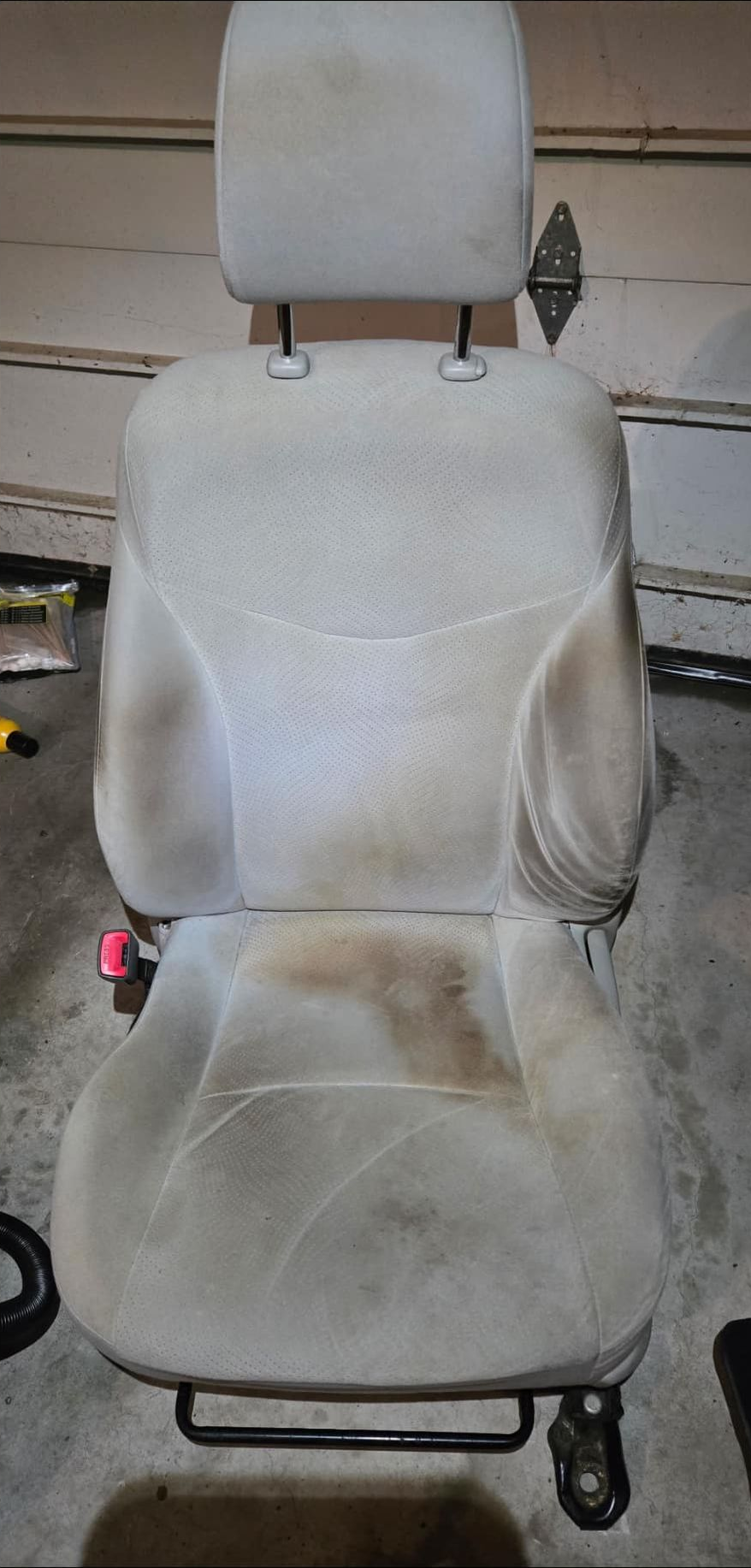 A heavily stained tan car seat with a headrest, set in a garage or workshop.