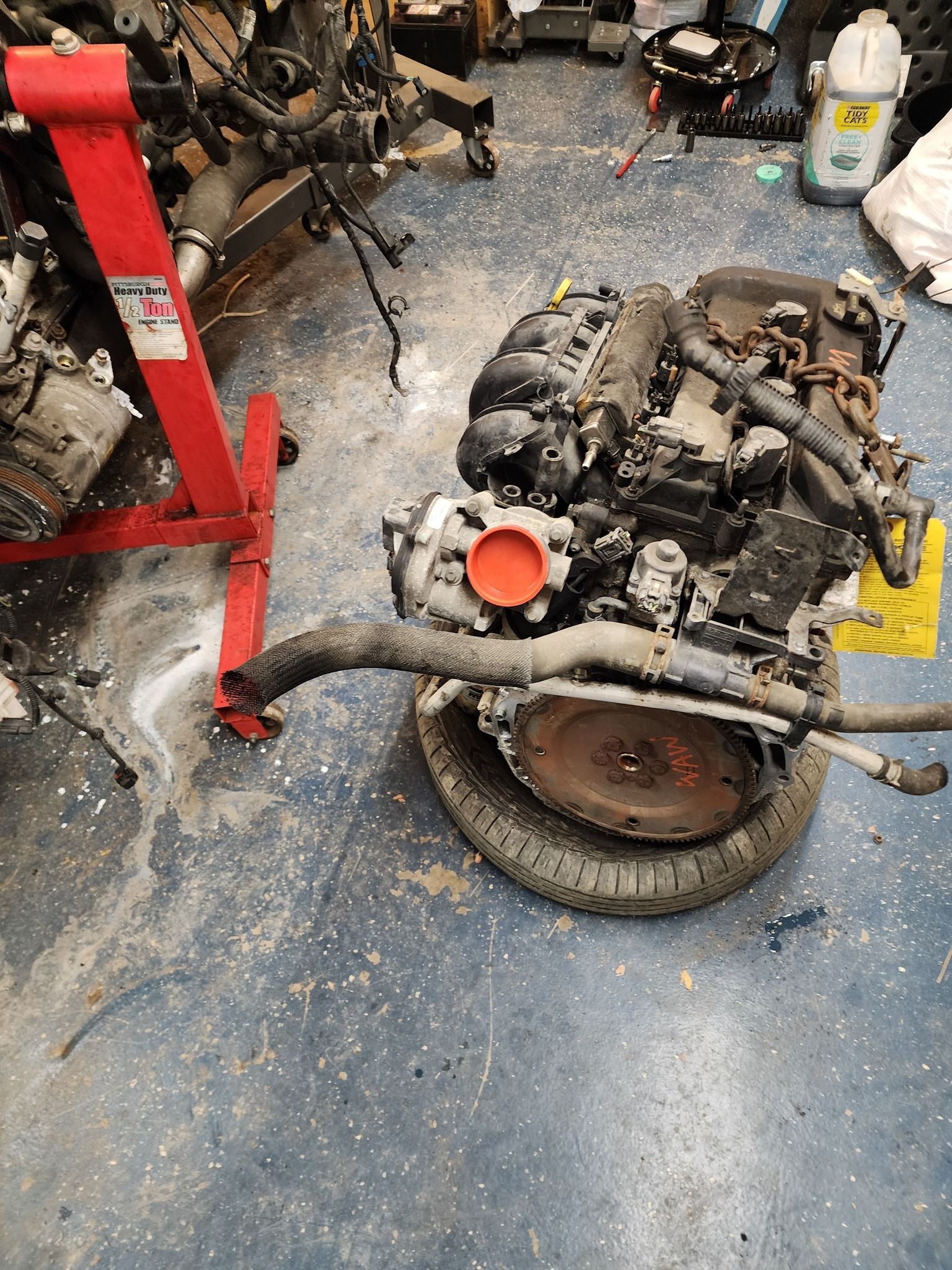 Engine on a tire in a cluttered workshop, near an engine stand.