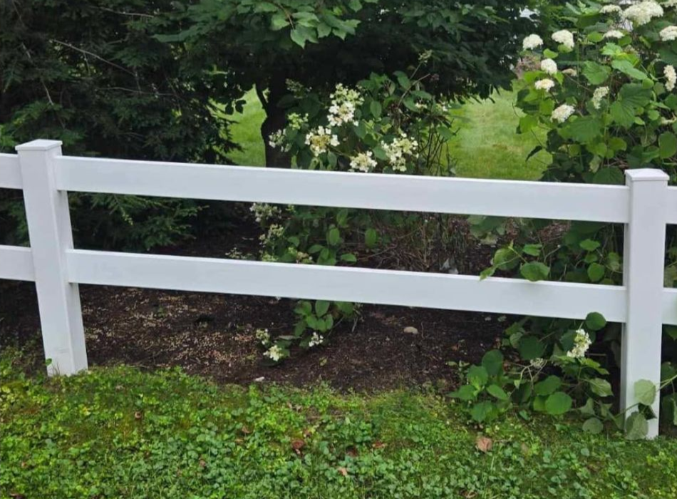 White vinyl fence in front of shrubs and green grass, with a blurred background.