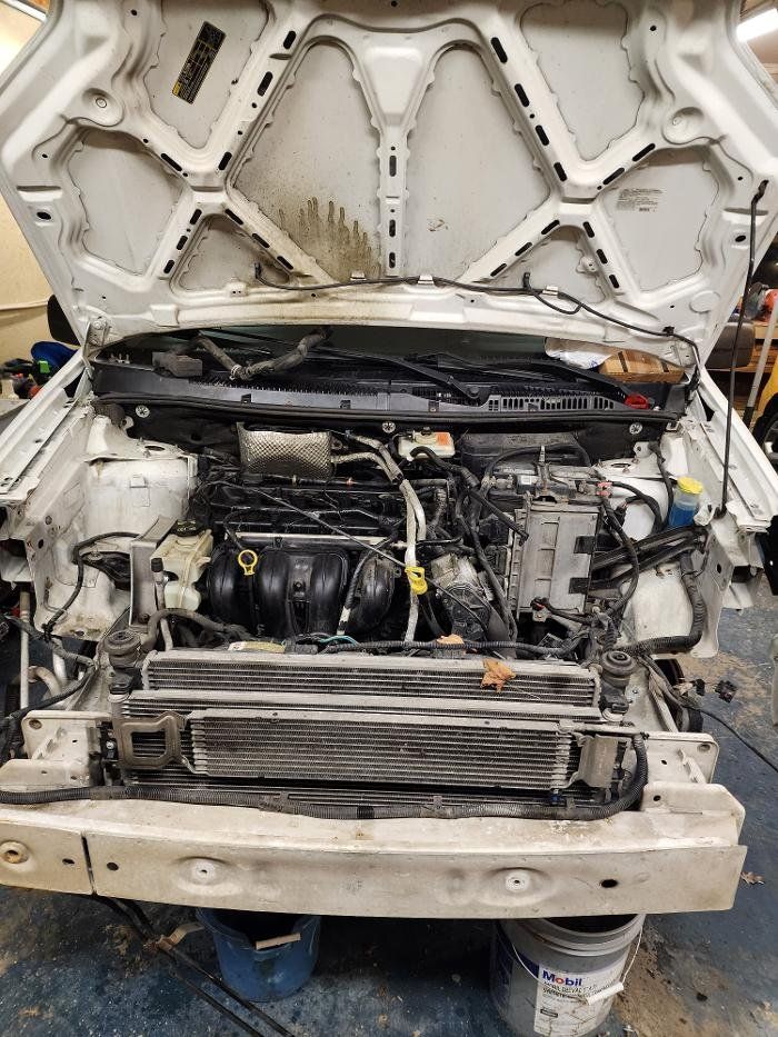 Car engine bay, open hood, white body, mechanical parts exposed, dirty, industrial setting.