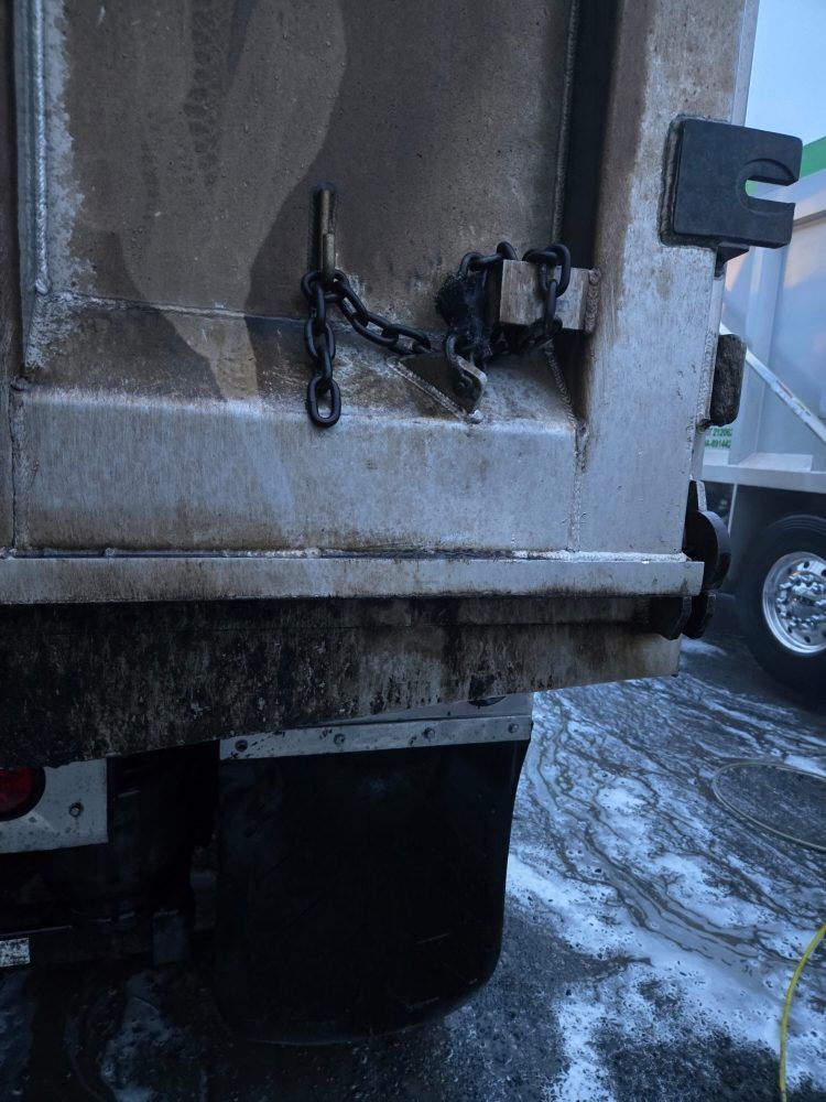 Back of a dump truck with the tailgate secured by a chain. Black mud flap and part of a tire visible.