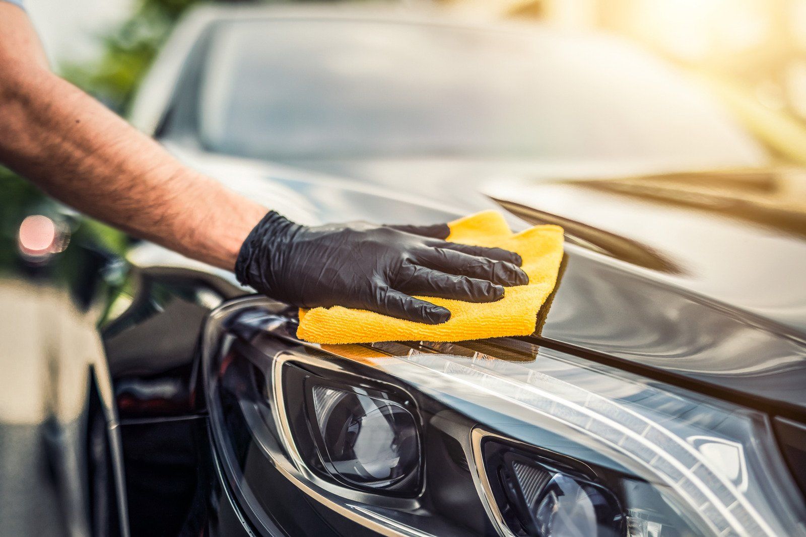 Person wearing black gloves cleaning a black car's hood with a yellow cloth in sunlight.