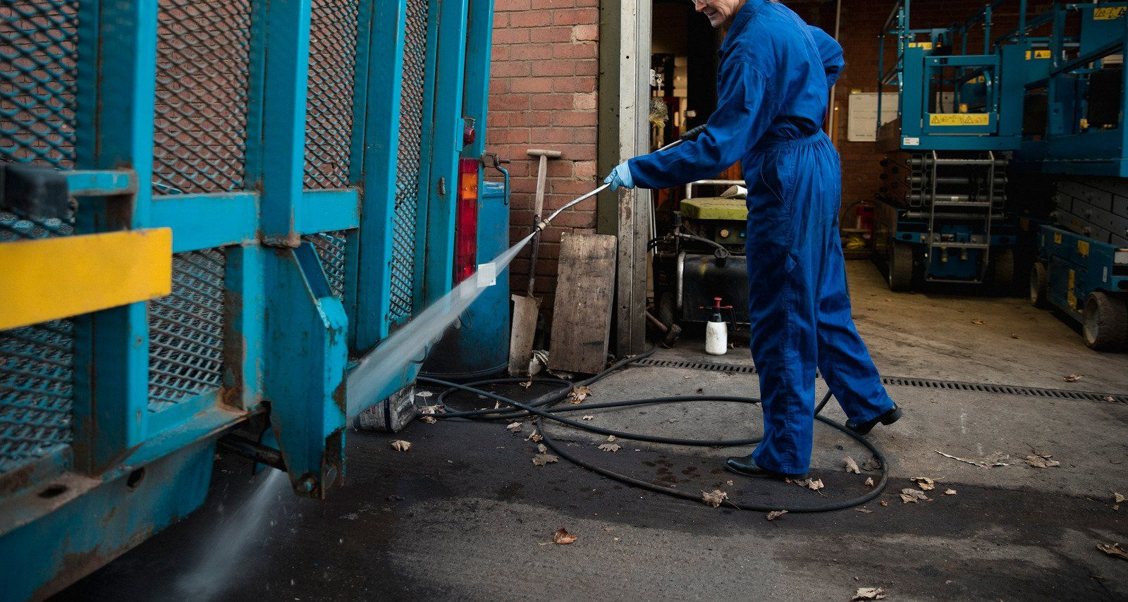 Person in blue overalls uses a pressure washer to clean a large blue structure in a warehouse.