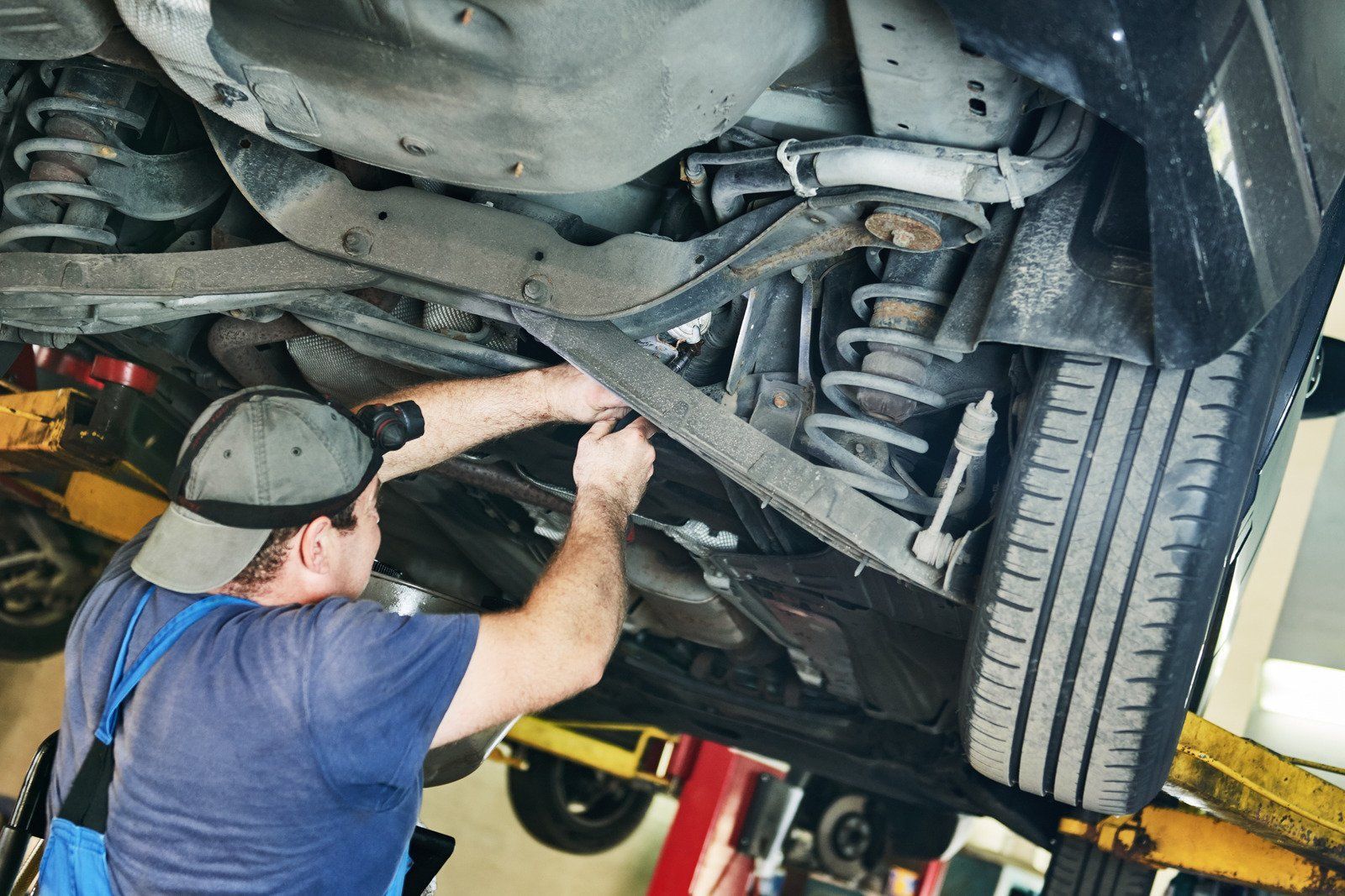 Mechanic working under a car on a lift. He is wearing a hat and blue shirt.