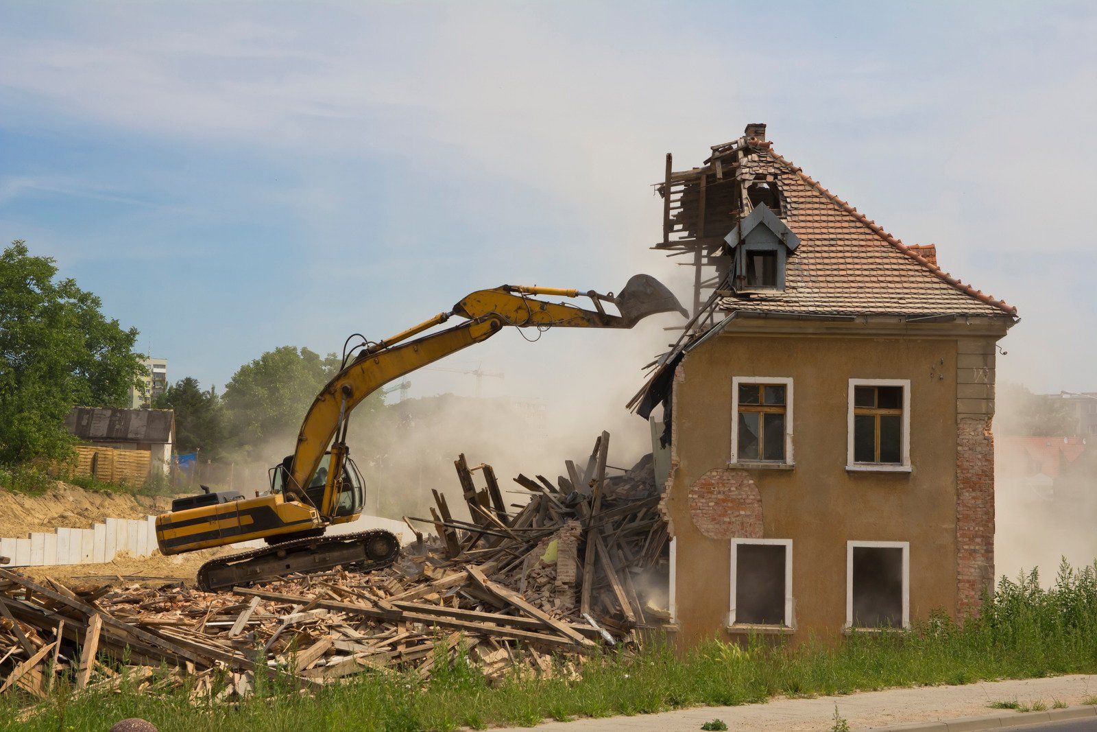 An excavator demolishes a two-story building, clouds of dust billowing, on a sunny day.