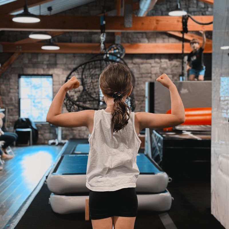 Person flexing arms in a gym in Gibbsboro, NJ, seen from behind near workout equipment and hanging rings