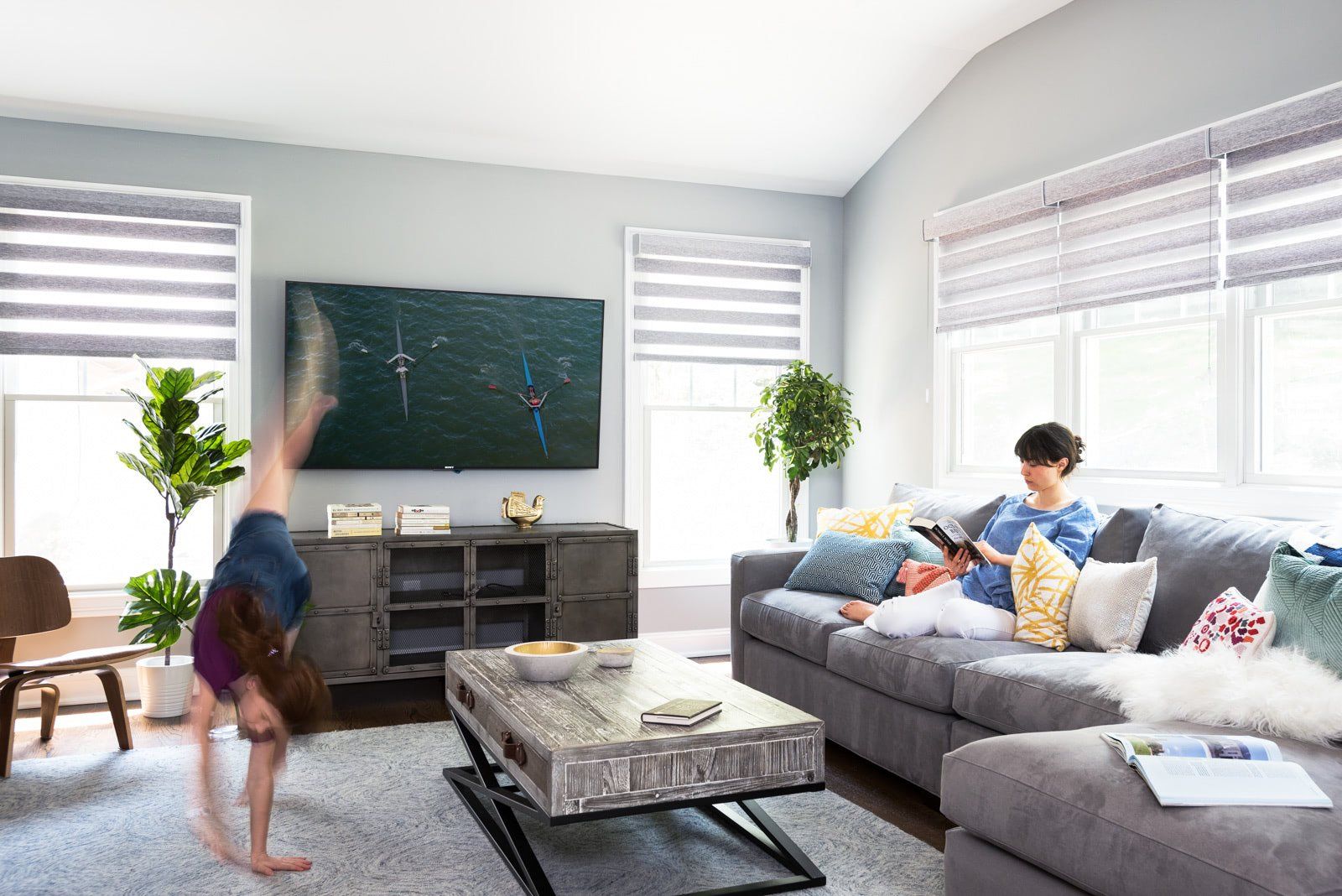 A woman is reading a book while a child does a handstand in a living room.