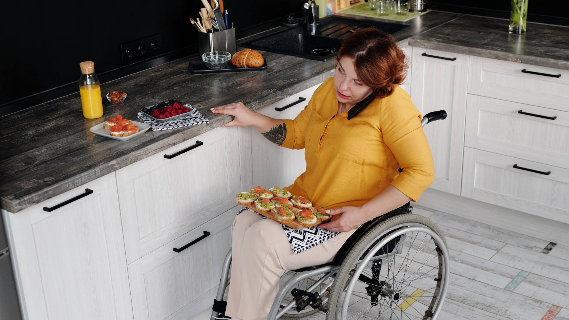 A woman in a wheelchair is sitting in a kitchen holding a tray of food.
