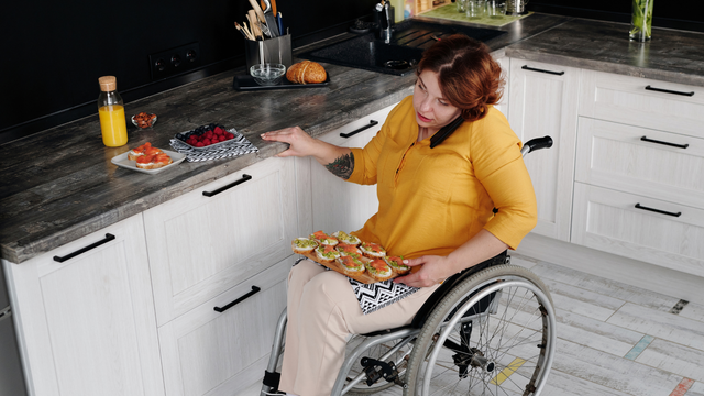 A woman in a wheelchair is sitting in a kitchen holding a tray of food.