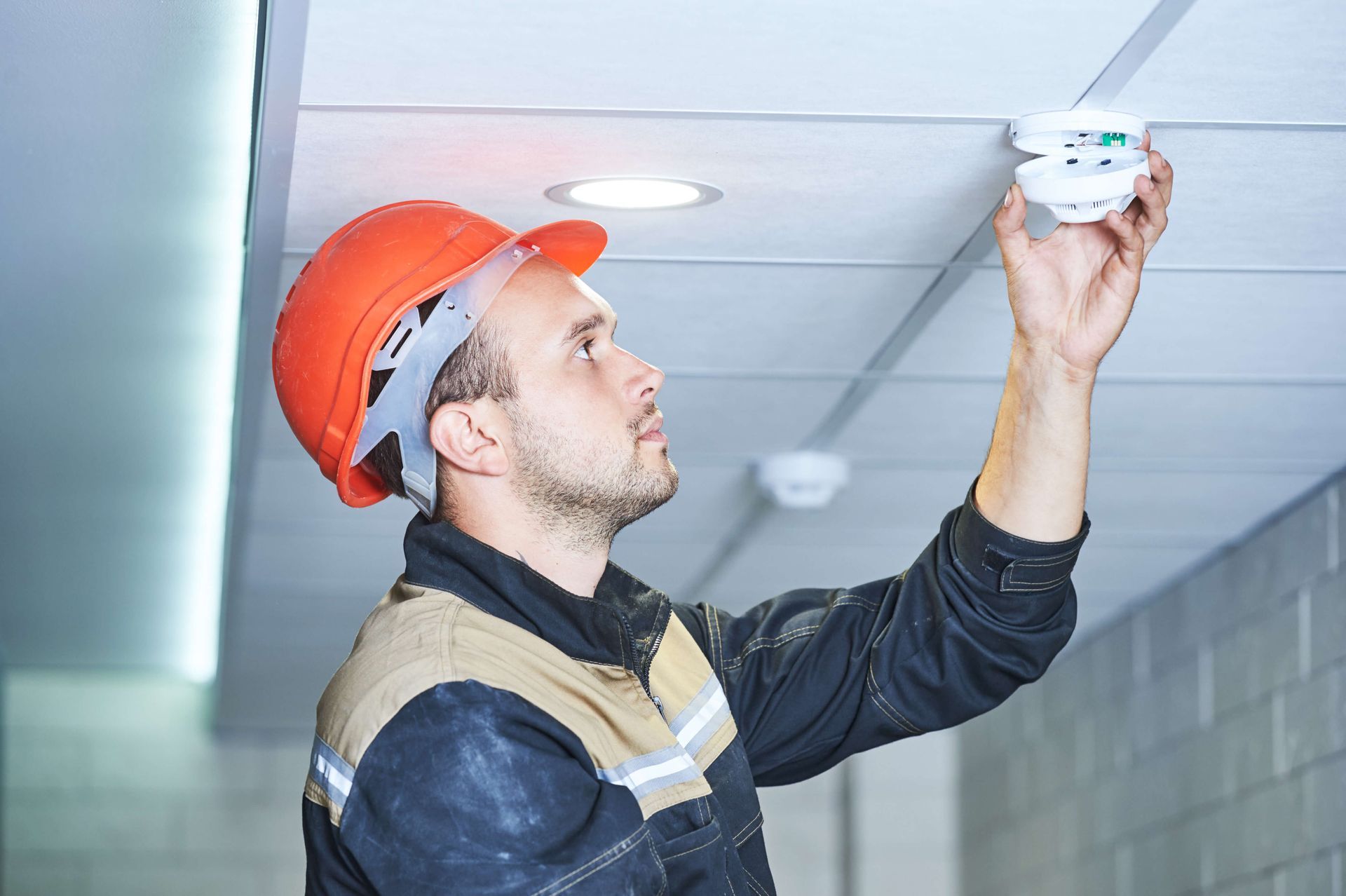 A man in a hard hat is installing a smoke detector on the ceiling.