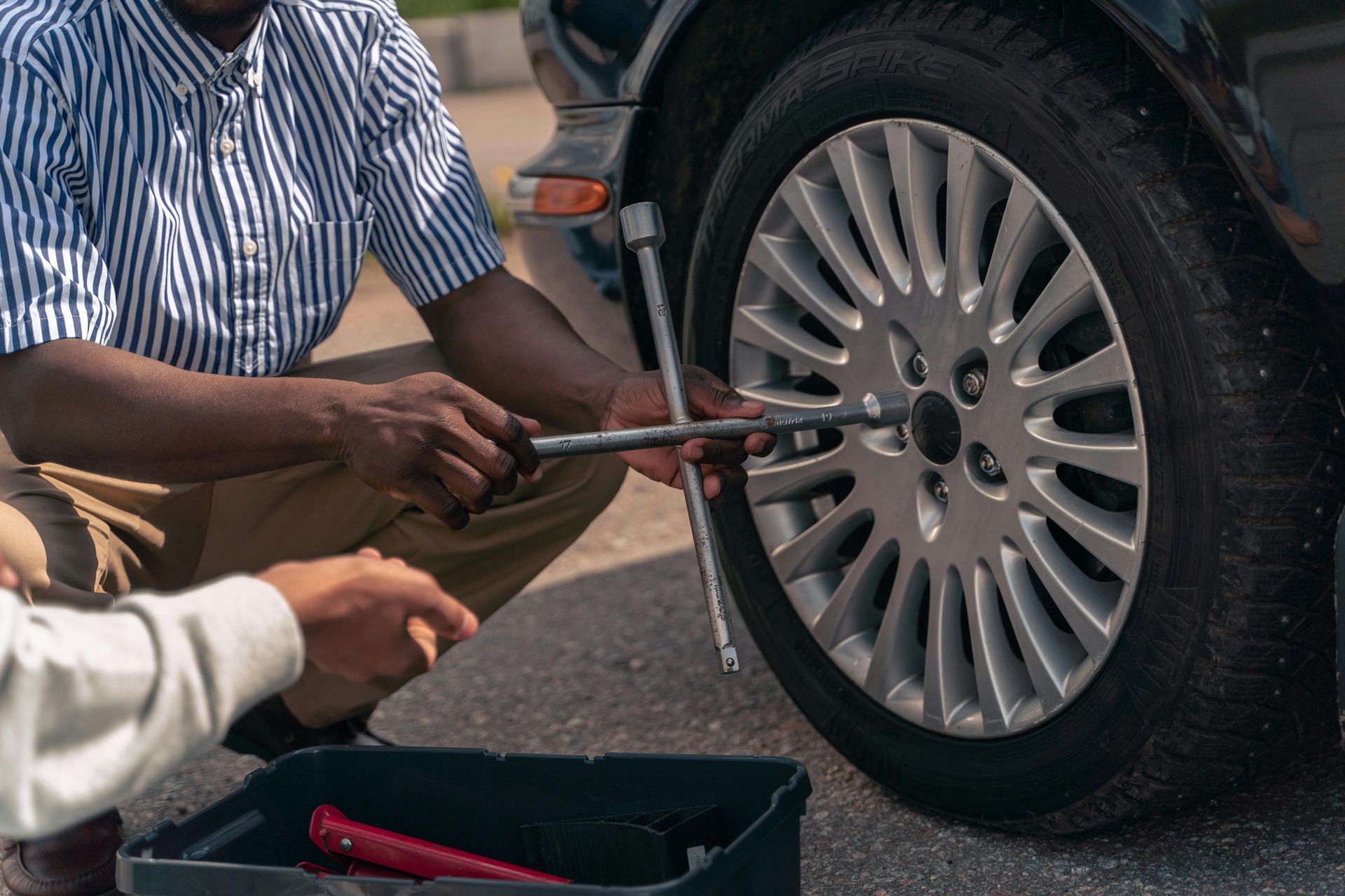 Person kneeling beside a car, using a lug wrench to remove a wheel. Another person holds a black toolbox.