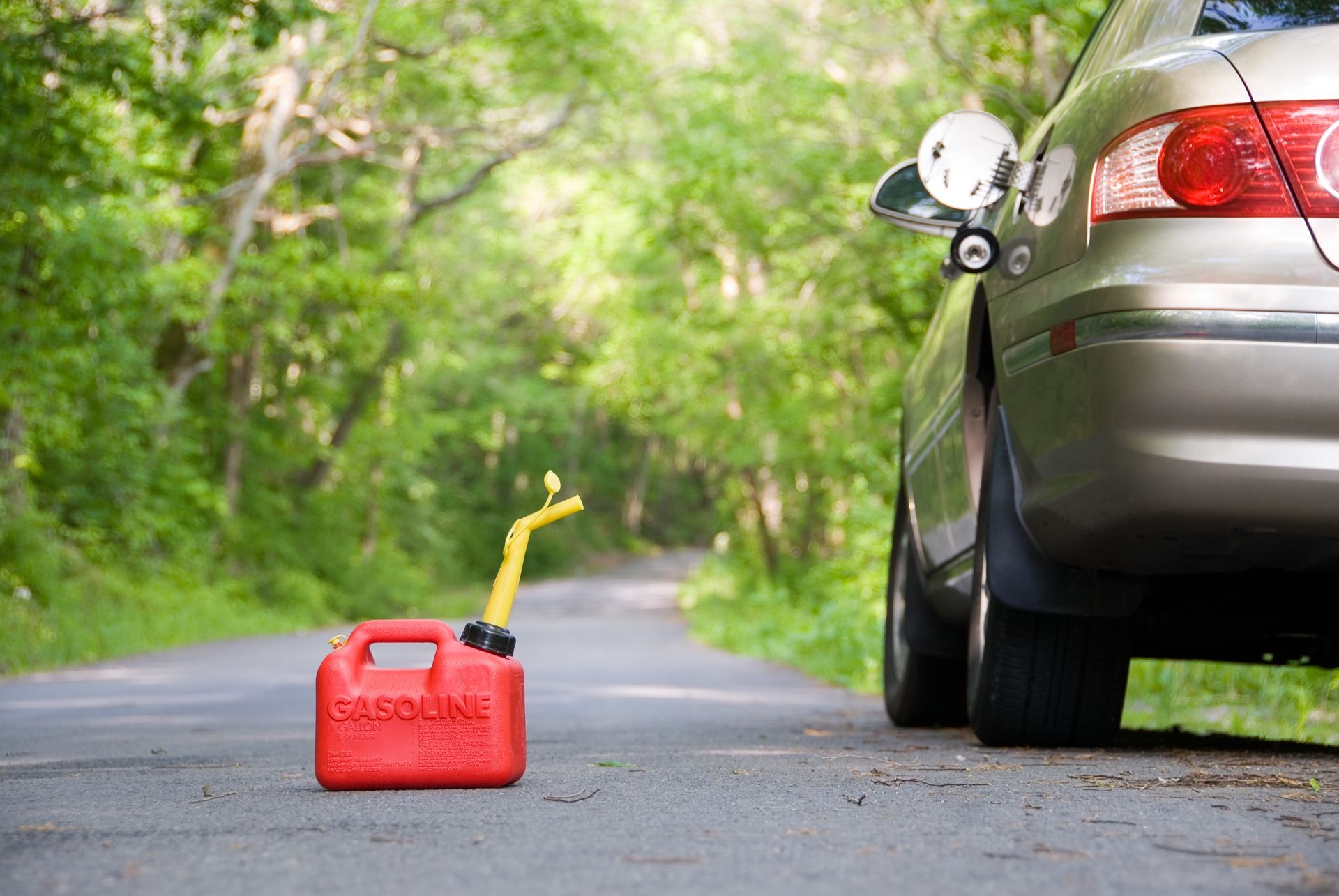 Red gas can on a road, with fuel nozzle raised. A car is parked nearby in a green, wooded area.