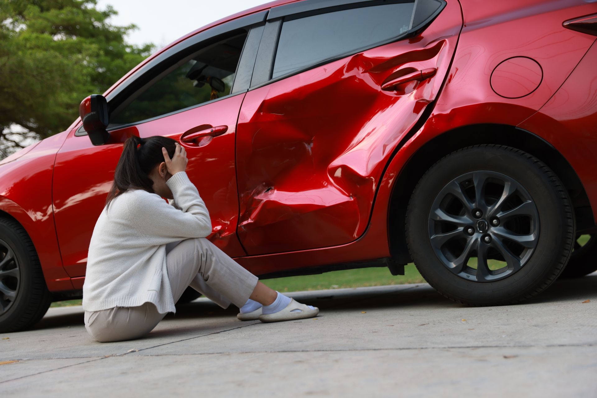 Woman sits beside a damaged red car, holding her head in distress.