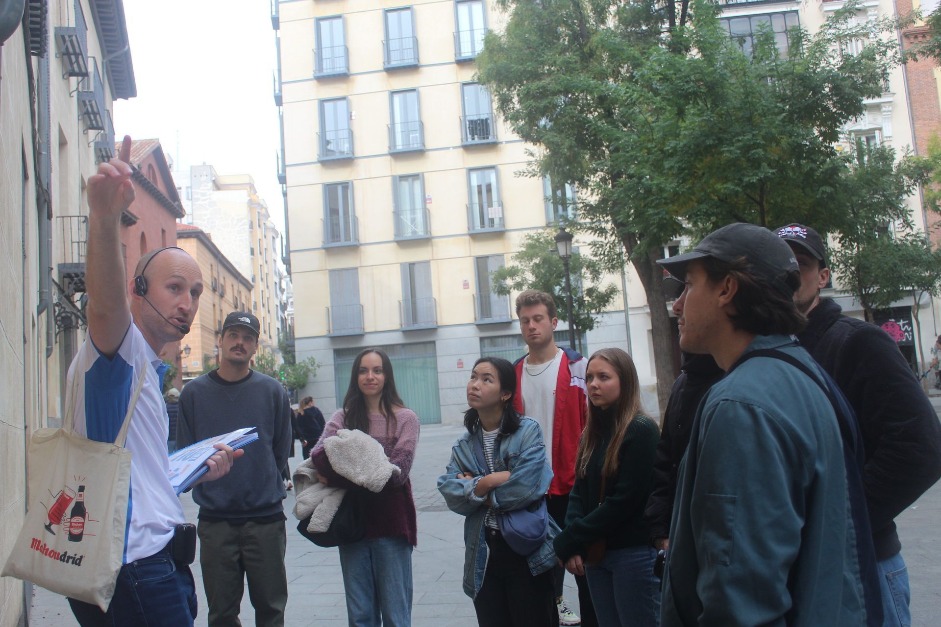 A Madrid Free Walking Tour guide gestures enthusiastically while speaking to a group of tourists in a tree-lined plaza.