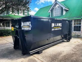 A dumpster is parked in front of a house with a green roof.