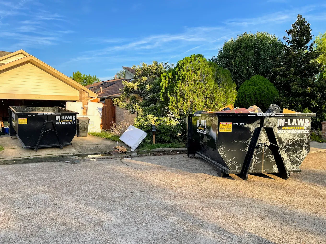 Two black dumpsters on a driveway with a house and trees in the background under a blue sky.