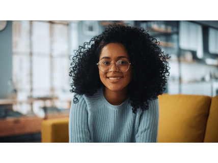 A woman with curly hair and glasses is sitting on a couch and smiling.