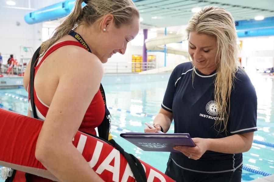 Two women at an indoor pool, one holding a tablet while the other leans in to look at it.