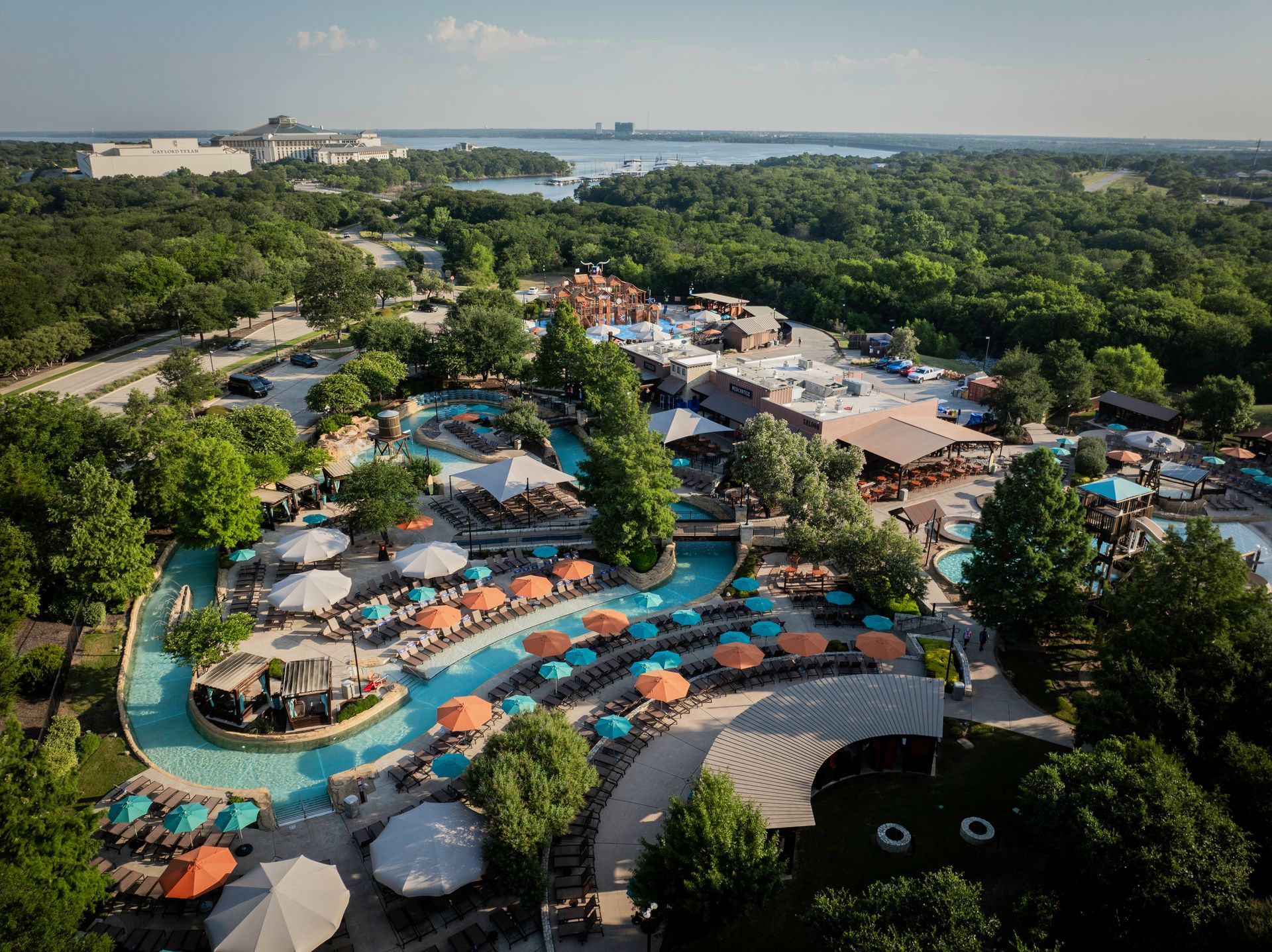 Aerial view of a tropical resort with winding pools, teal umbrellas, palm trees, and the ocean beyond.