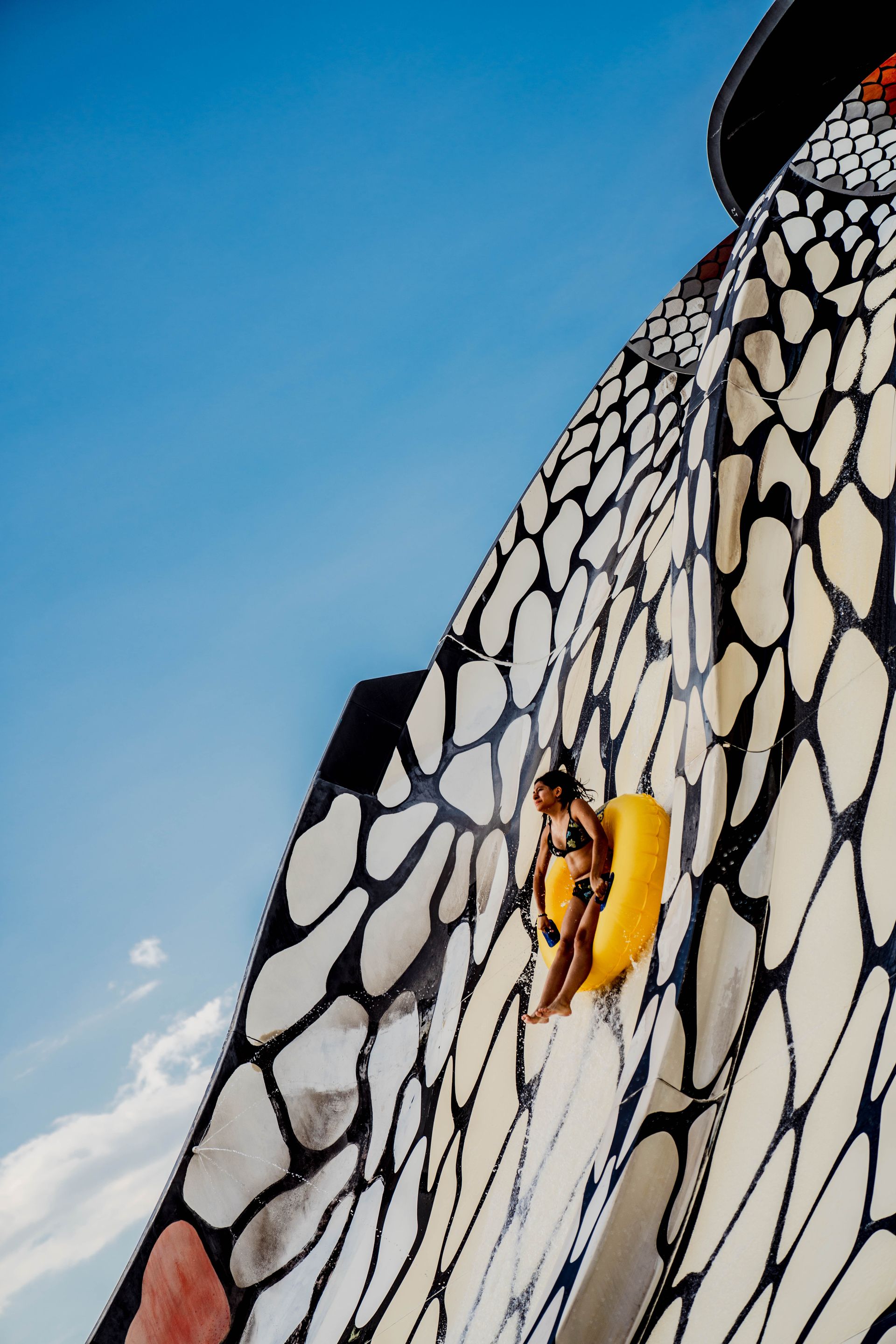Yellow kayaker climbing a towering black-and-white patterned wall under a blue sky