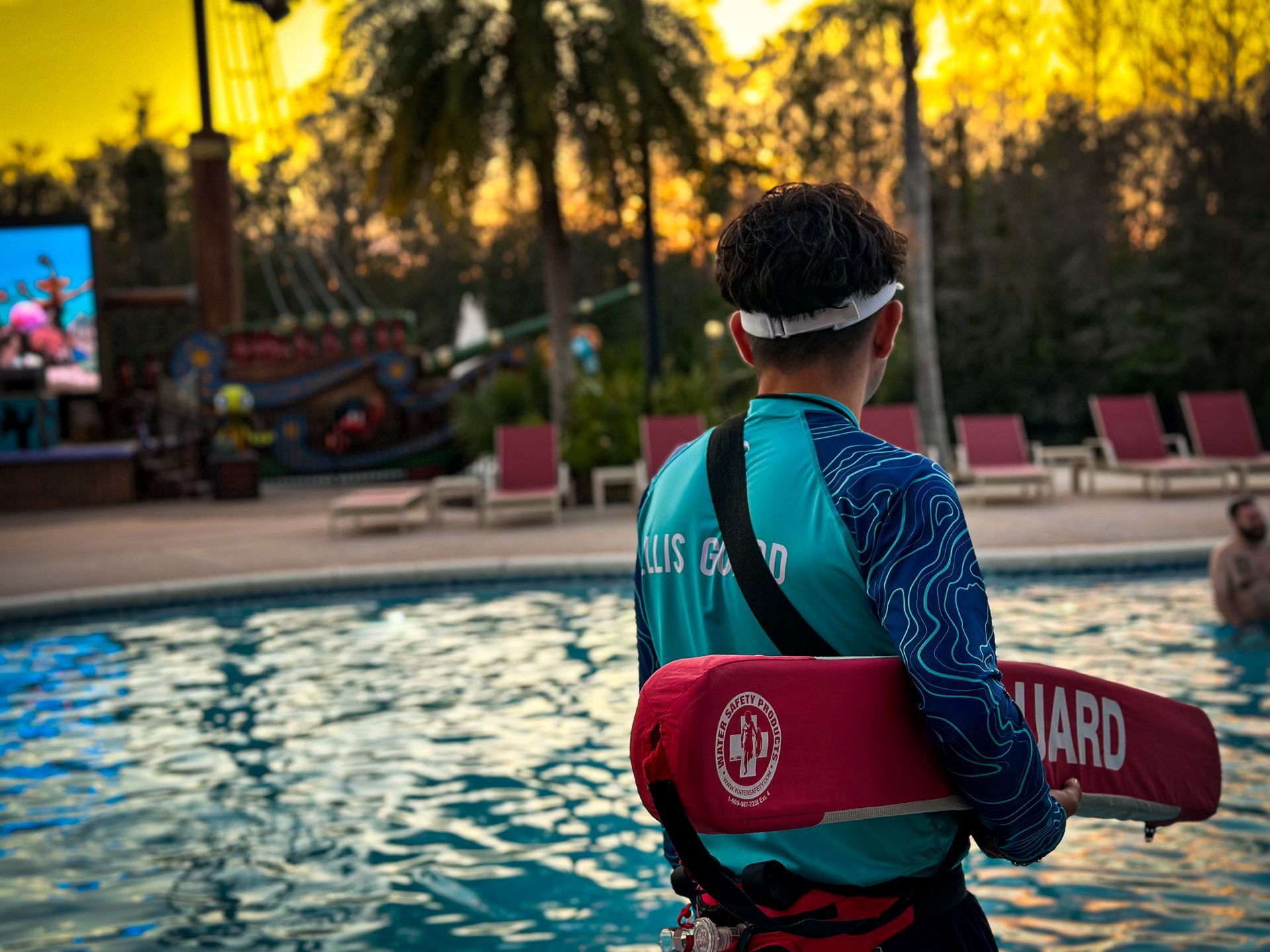 Lifeguard standing by a pool at sunset, holding a red rescue float