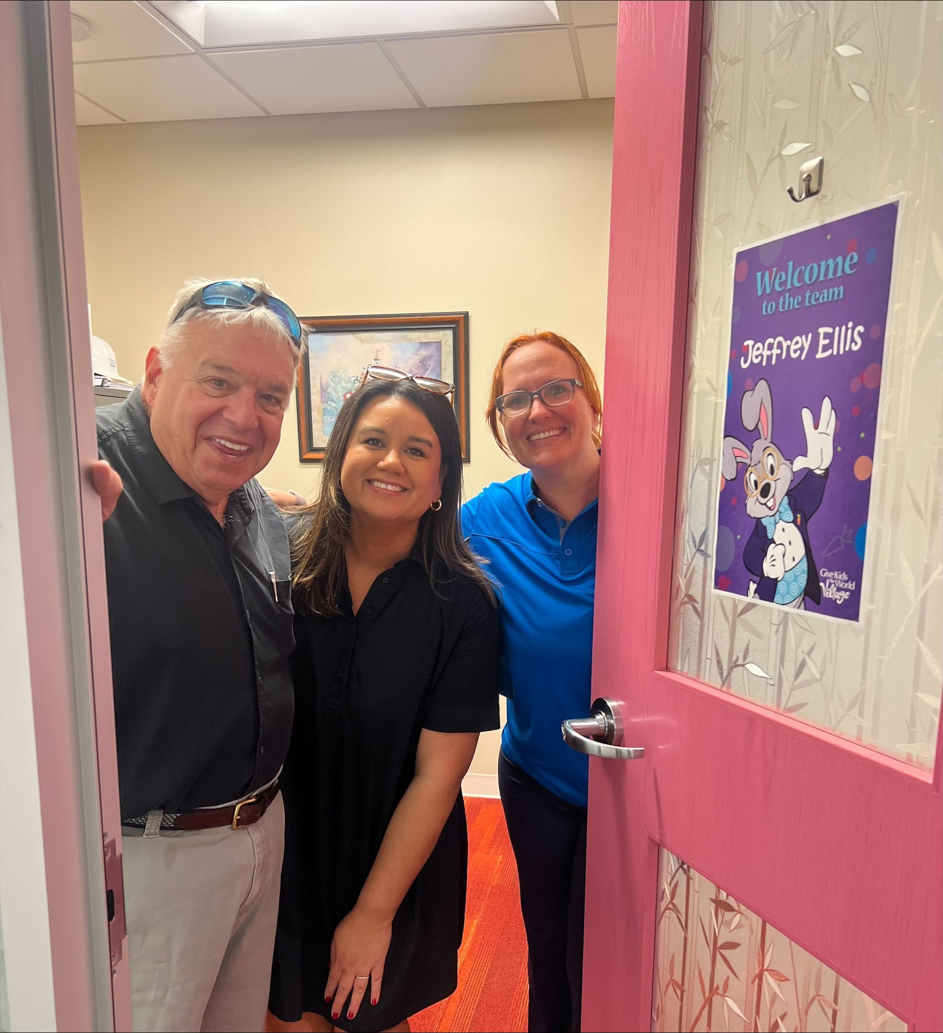 Three people smiling in a pink doorway beside a sign that says “Happy Easter.”