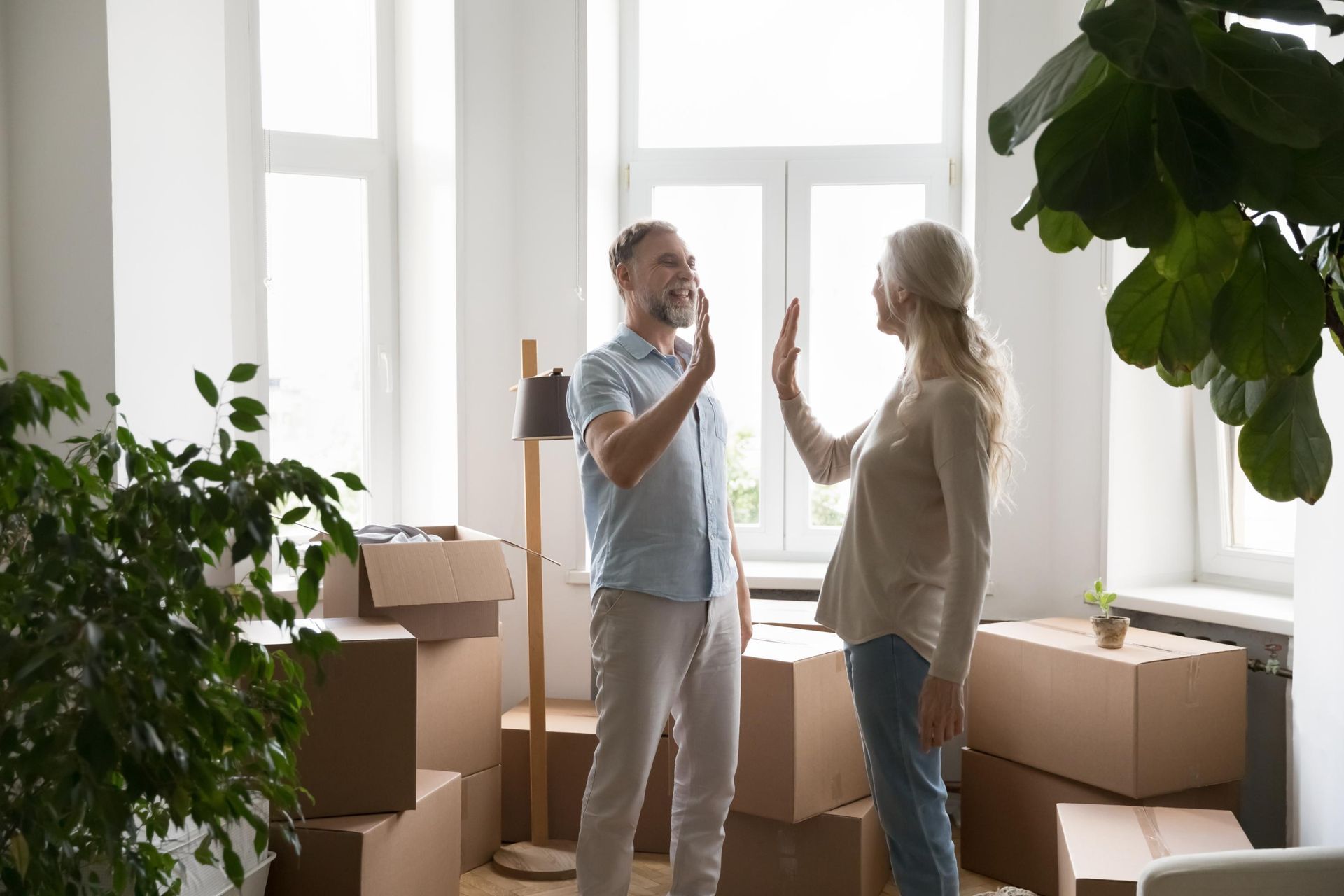 Couple high-fiving in a room with packed moving boxes and a window; celebrating a new home.