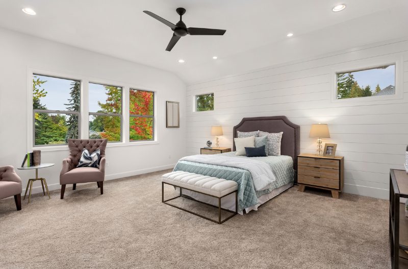 A carpeted bedroom with a bed , chairs , nightstands and a ceiling fan.