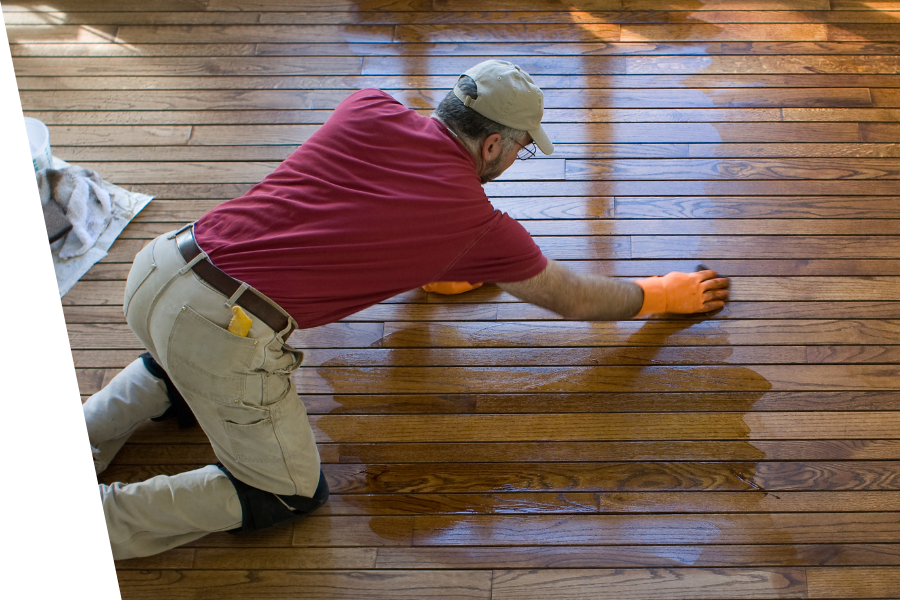 A man is kneeling on the floor refinishing a wooden floor.