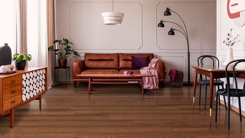 A cozy living room featuring rich wooden flooring, a brown leather sofa, a unique wooden sideboard with geometric patterns, and modern decor elements like hanging lamps, plants, and contemporary artwork on the wall. A small dining area with stylish chairs is visible to the right.