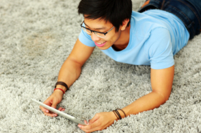 man in a blue shirt lying on the carpet while using his tablet
