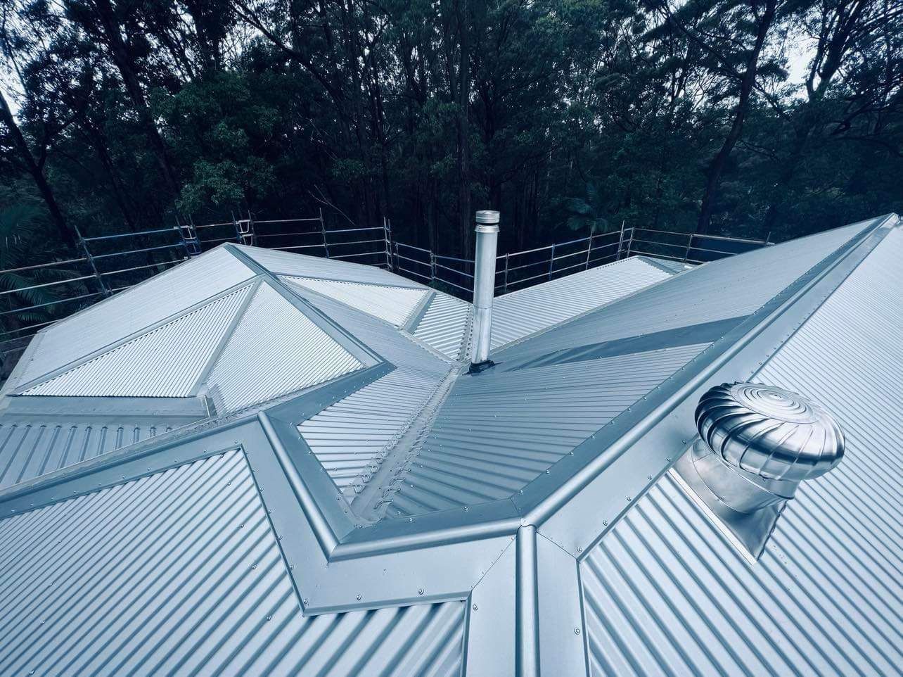 A Metal Roof With a Chimney and Vent, Set Against a Backdrop of Green Trees — Venom Roofing Pty Ltd in Sunshine Coast, QLD