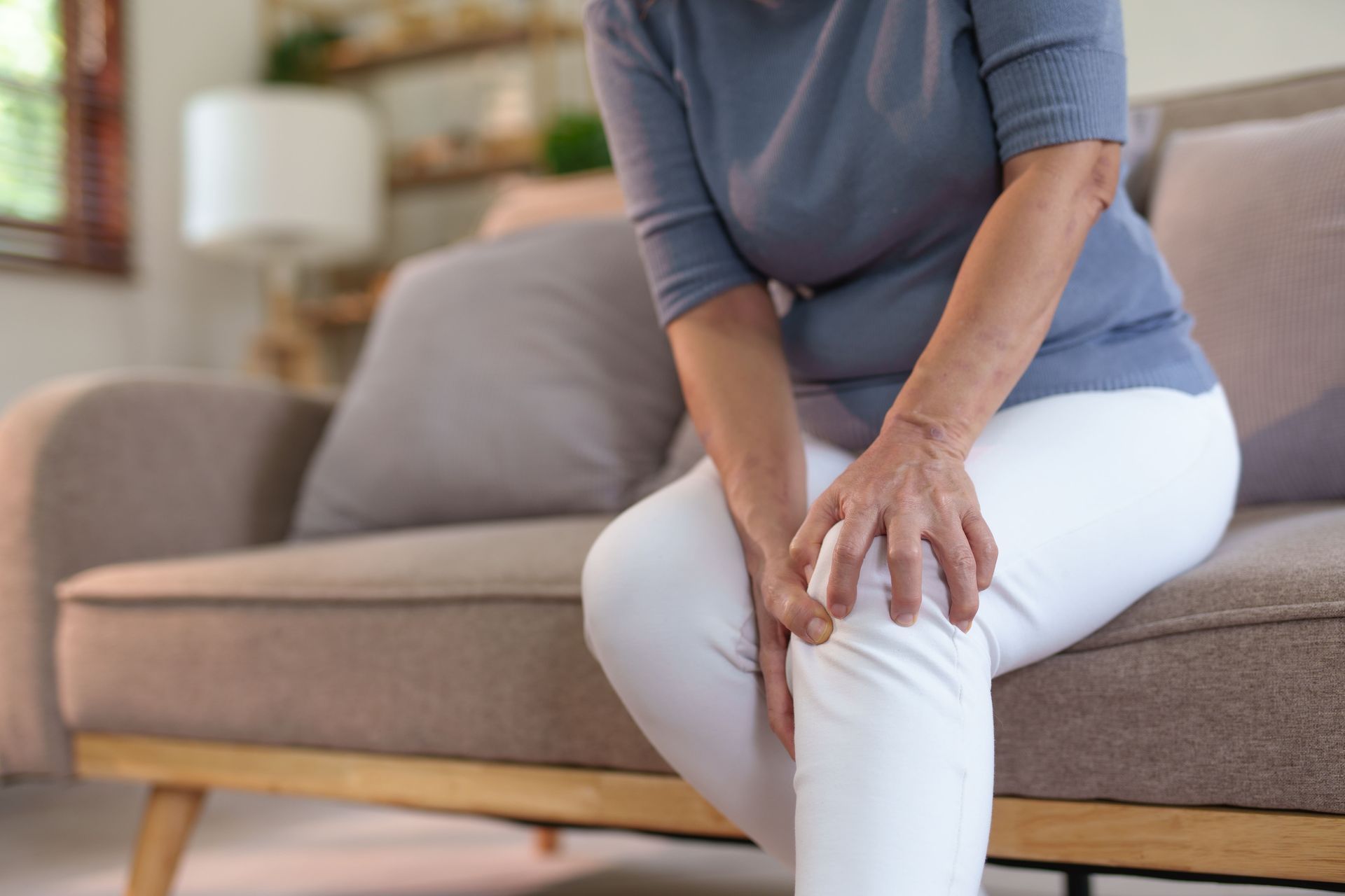 An elderly woman is sitting on a couch holding her knee in pain.