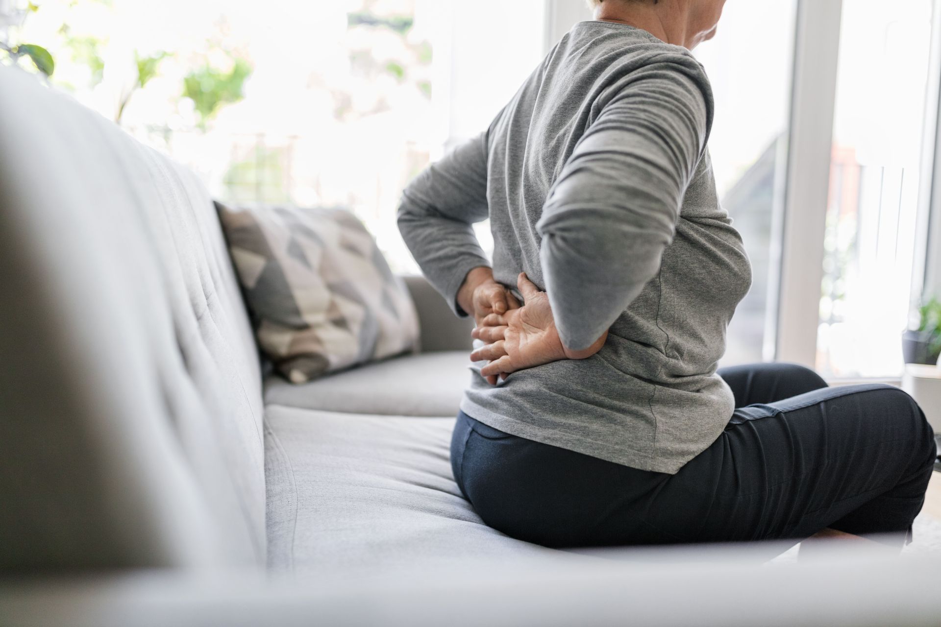 A woman is sitting on a couch holding her back in pain.