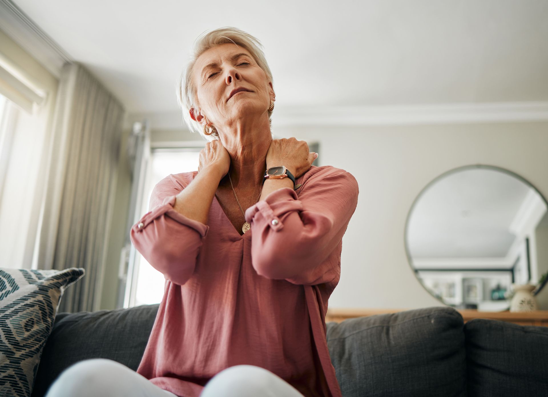 An elderly woman is sitting on a couch holding her neck in pain.