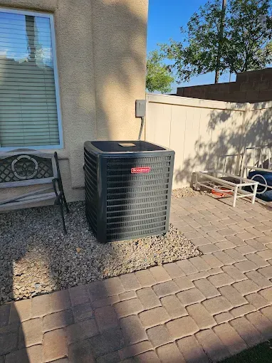 Air conditioning unit outside a building, set on gravel. A wooden fence and patio are in the background.