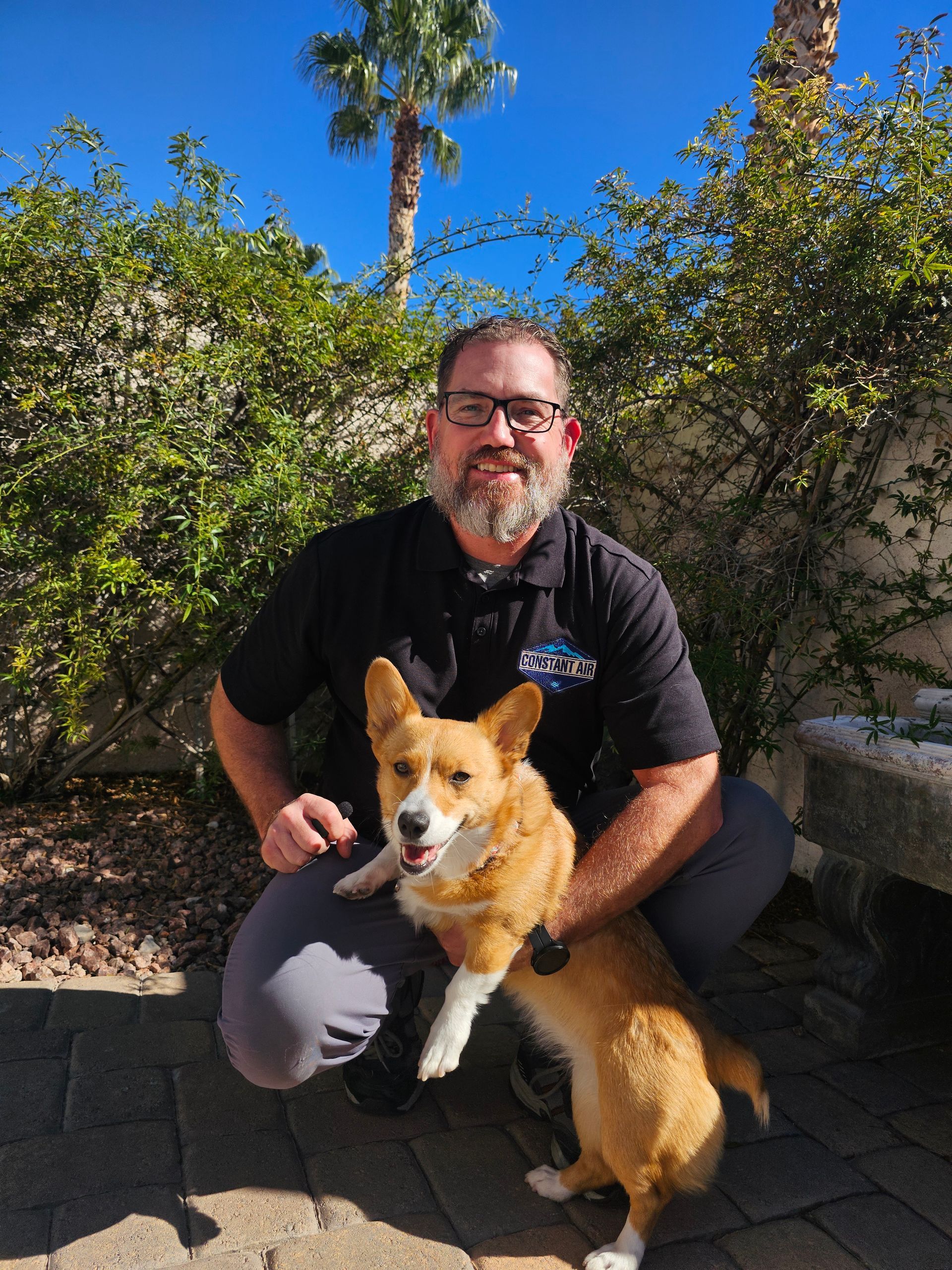 Man kneeling, holding a corgi dog in front of foliage. Sunny day.