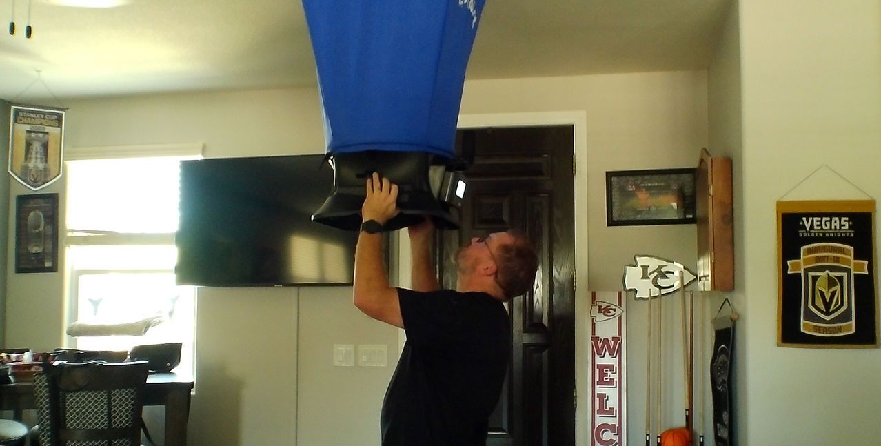Man holding a large blue and black object overhead indoors. Room with a TV and wall decor.
