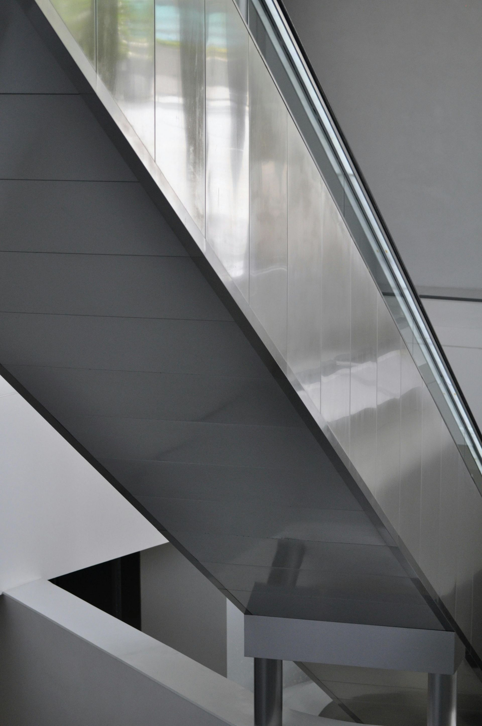 Modern, angled escalator with reflective silver metal paneling and glass railing in a brightly lit space.