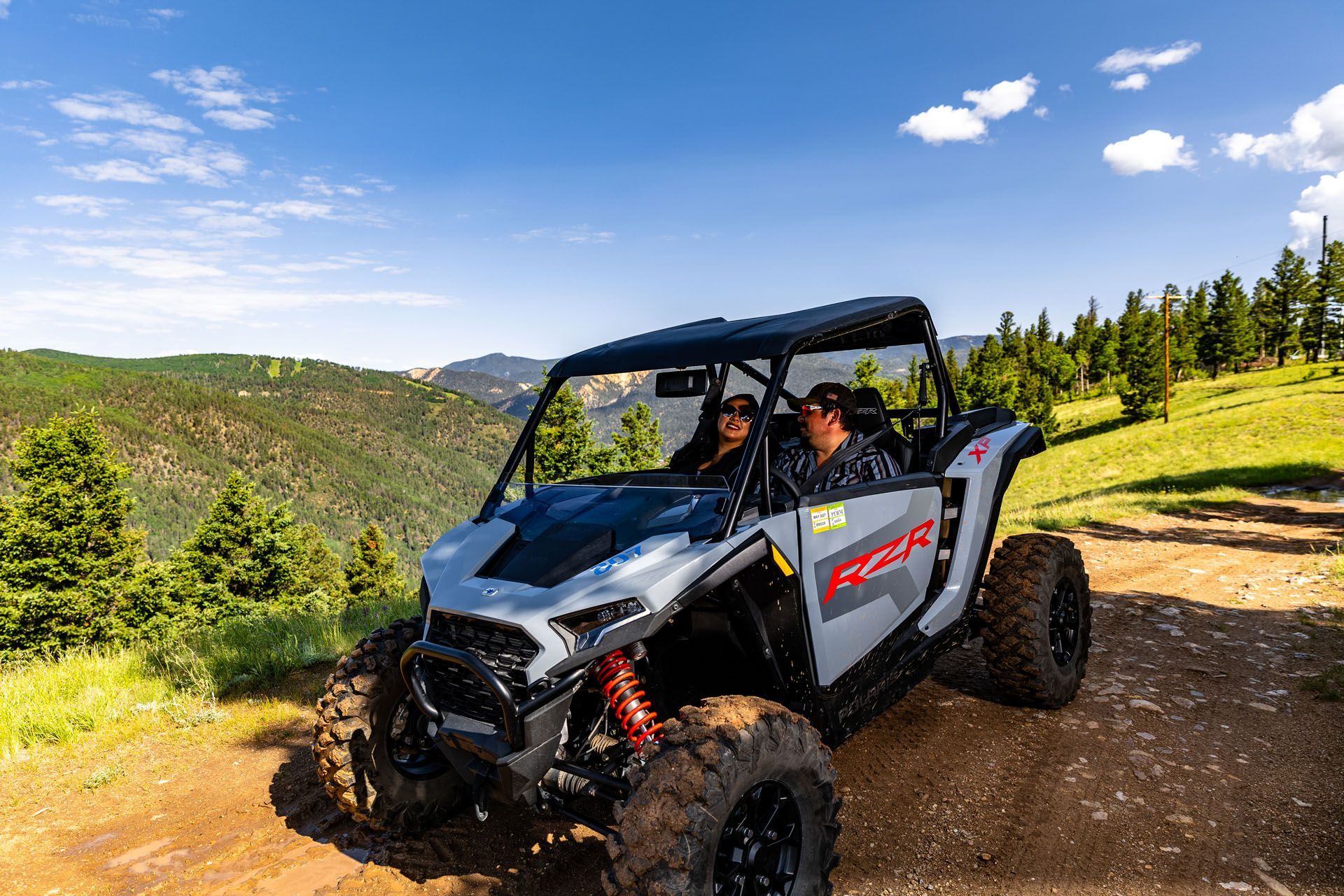 Gray UTV on a dirt road with two people, mountains in the background, and blue sky.