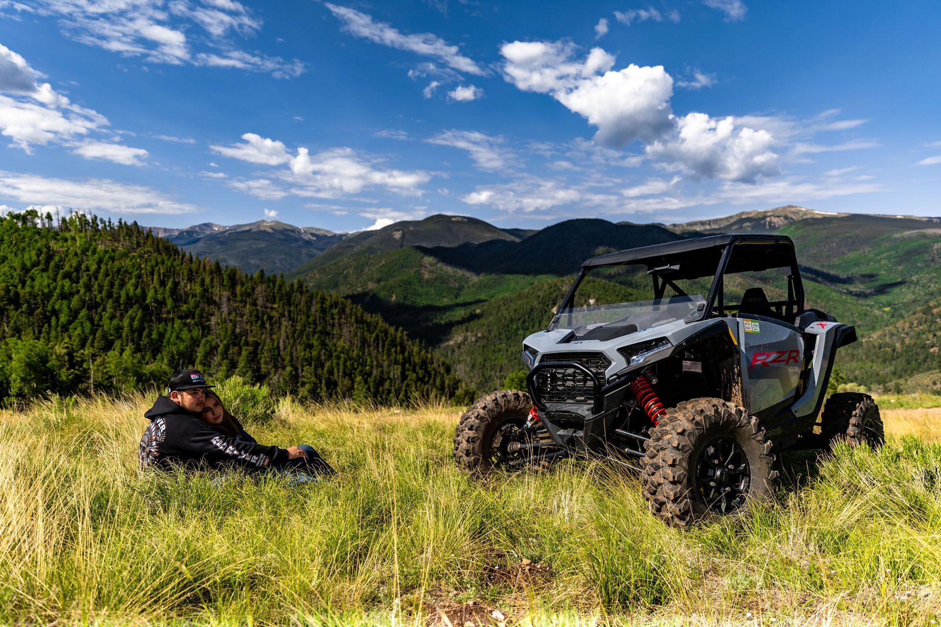 Side-by-side ATV parked on a grassy hill, a person resting nearby. Mountains in the background, blue sky.