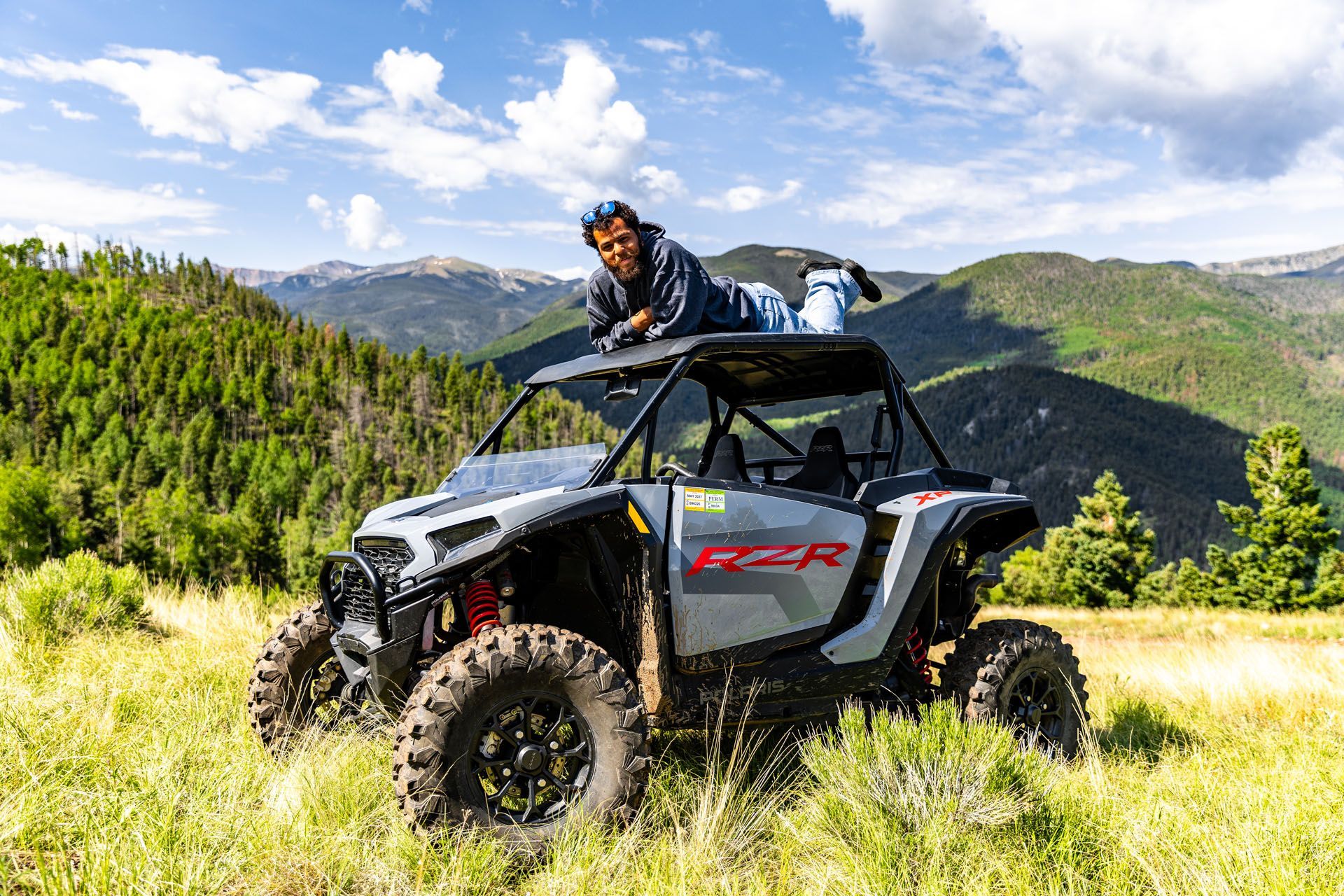 Man lying on the roof of a gray UTV in a grassy field with mountains in the background.