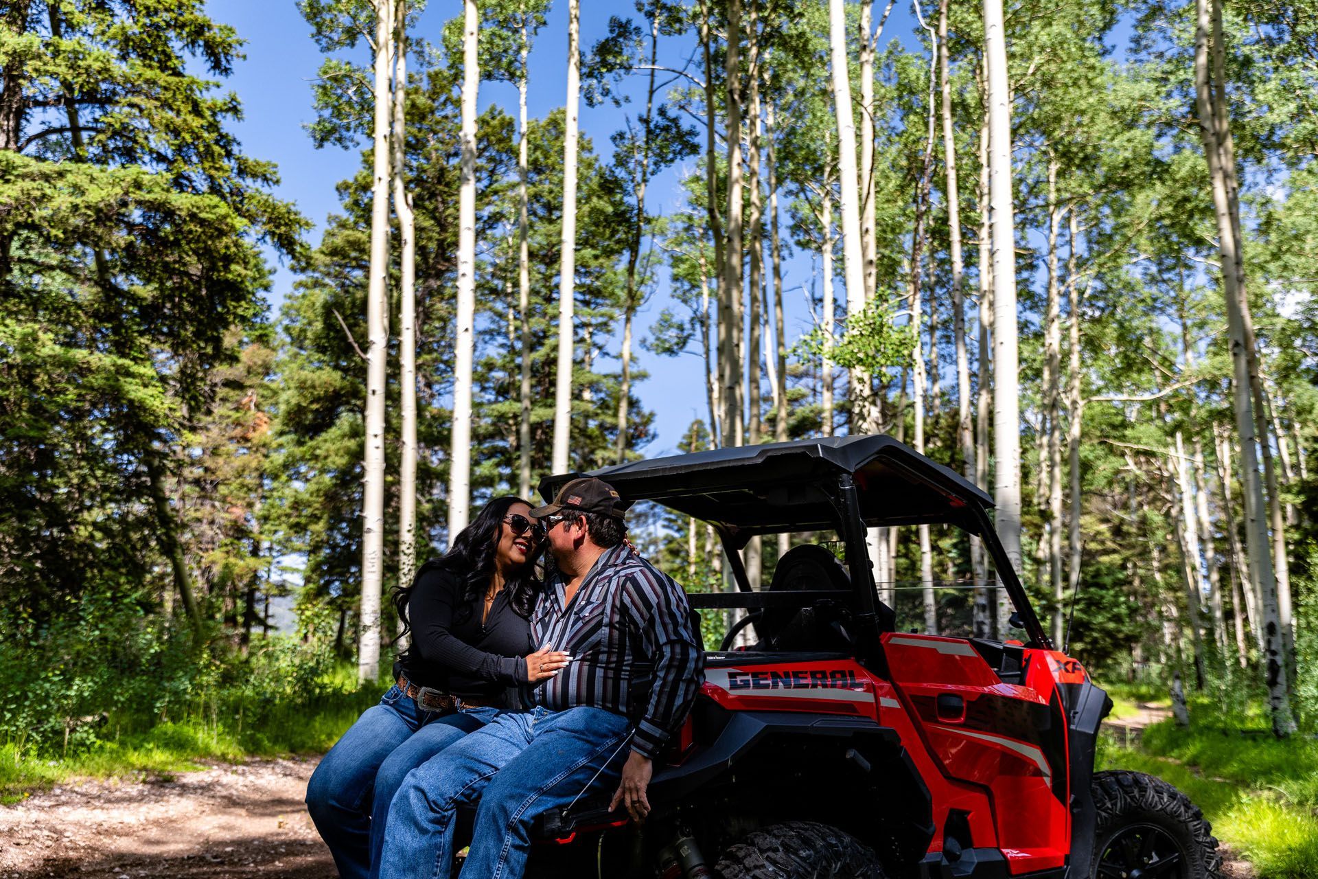 Couple kissing on a red ATV in a forest. Trees and blue sky in the background.