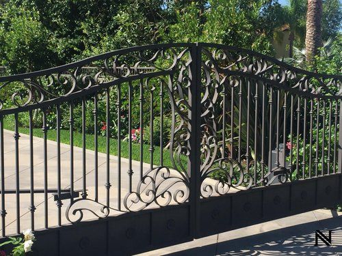 A black wrought iron gate is surrounded by trees and a driveway.