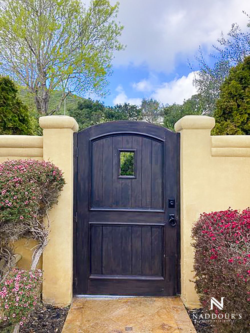 A wooden gate is surrounded by a yellow wall and flowers.