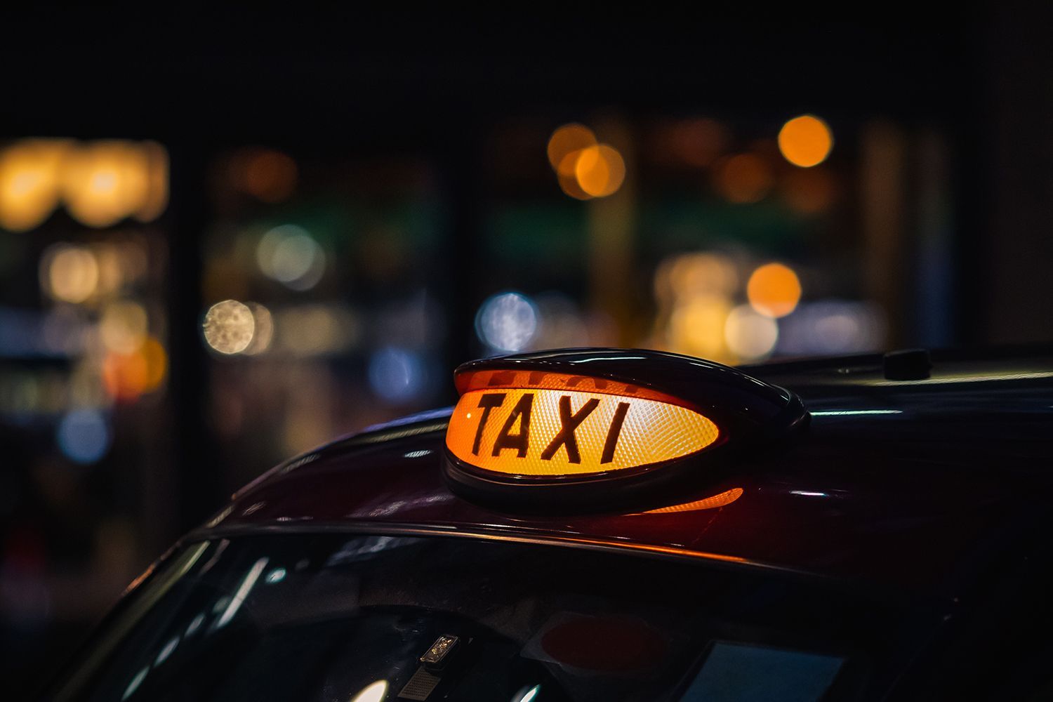 Illuminated taxi sign on a car roof at night with blurred city lights in the background.