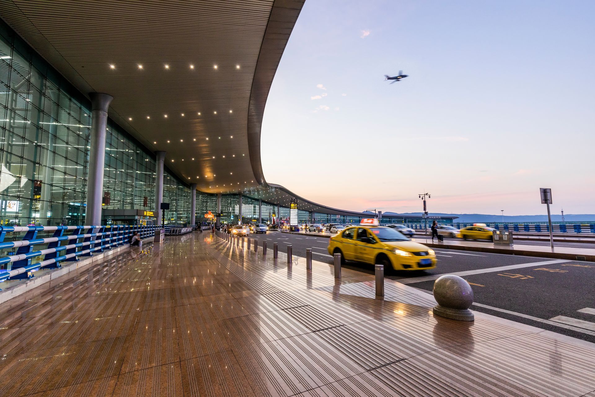 A yellow taxi is parked in front of an airport while a plane flies overhead.