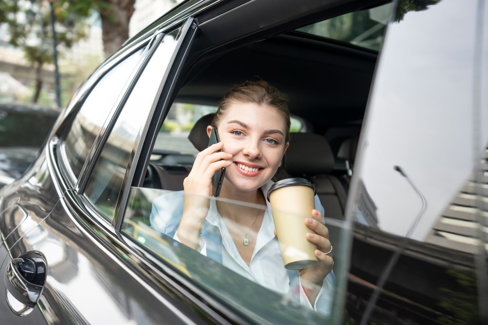 A woman is sitting in a car talking on a cell phone and holding a cup of coffee.